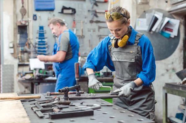 Werkstatt – junge Frau in Arbeitskleidung mit Augen- und Ohrenschutz arbeitet an einer Werkbank.