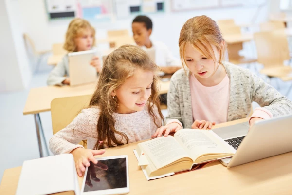 Schulklasse – zwei Mädchen schauen gemeinsam in ein Buch bzw. Computer